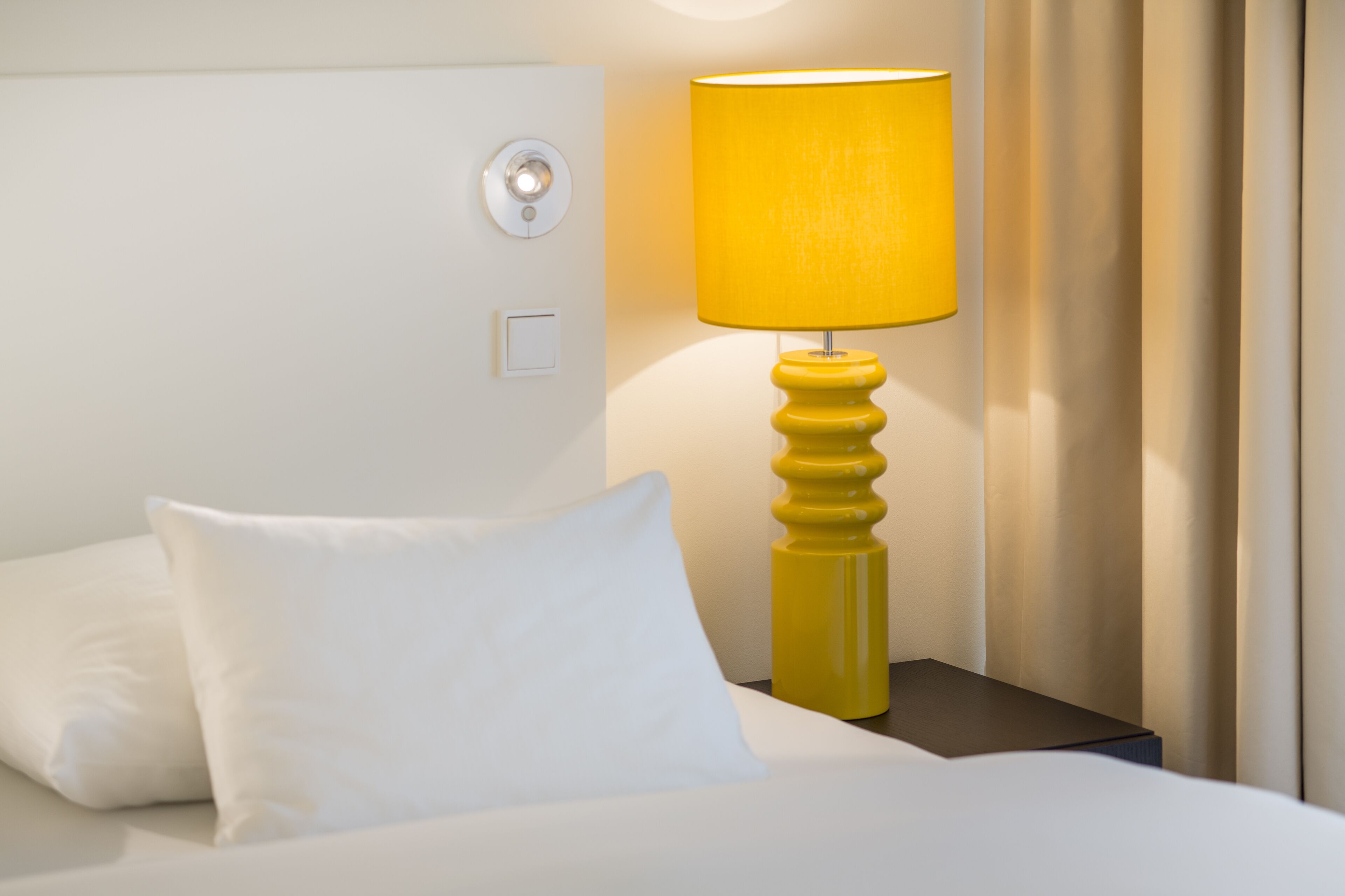 A neatly made bed with white bedding and pillows, a yellow lamp on a bedside table, and a wall switch.