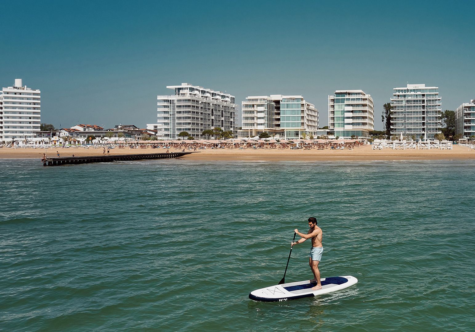 En person står på paddleboard på et roligt hav med en strand og moderne bygninger i baggrunden.
