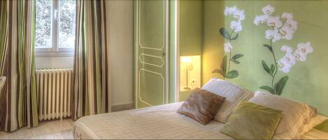 Un dormitorio con cama, almohadas, un radiador, cortinas y una pared con diseño floral.
