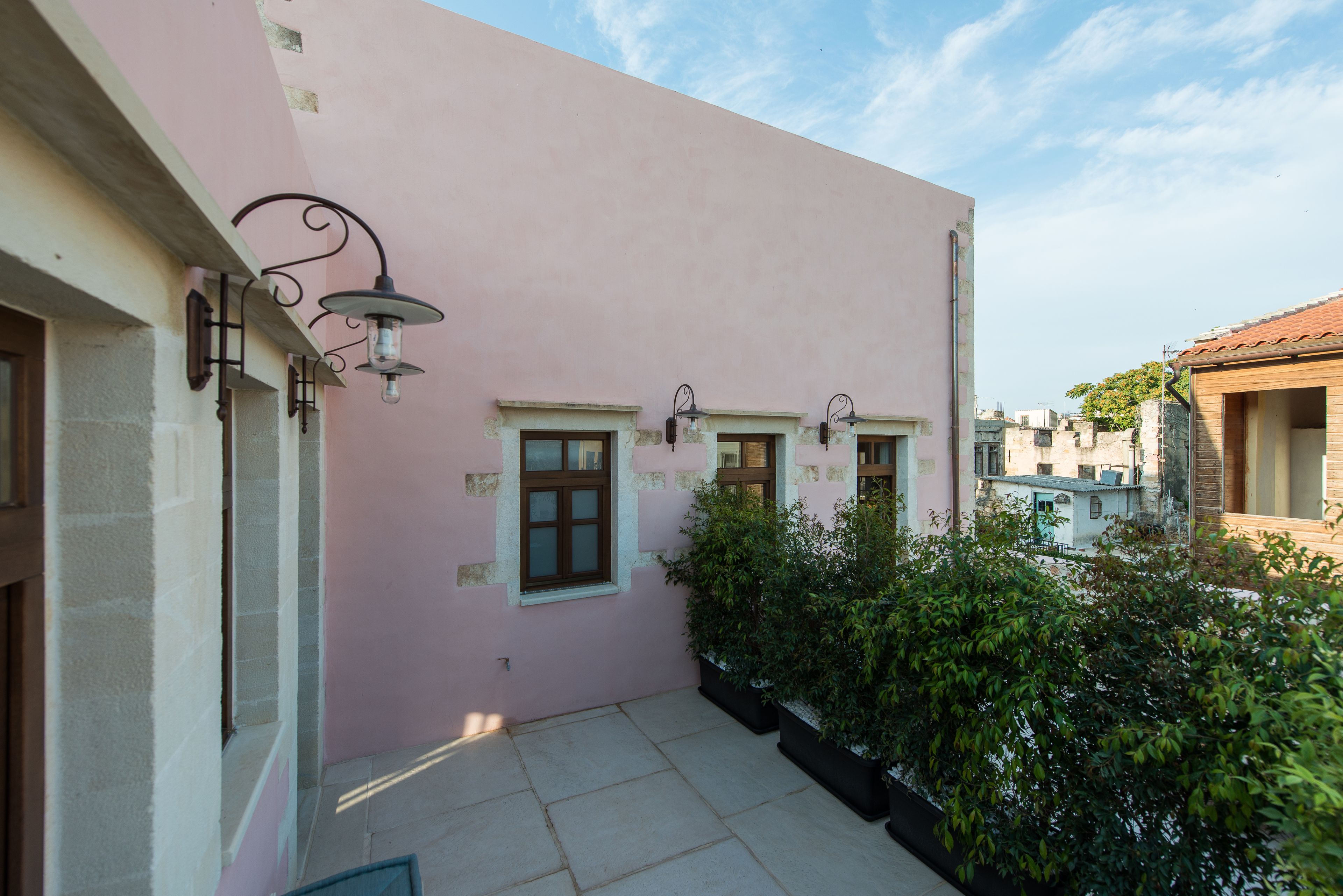 A pink building with two windows and a door, a small garden area with potted plants, and a clear sky.