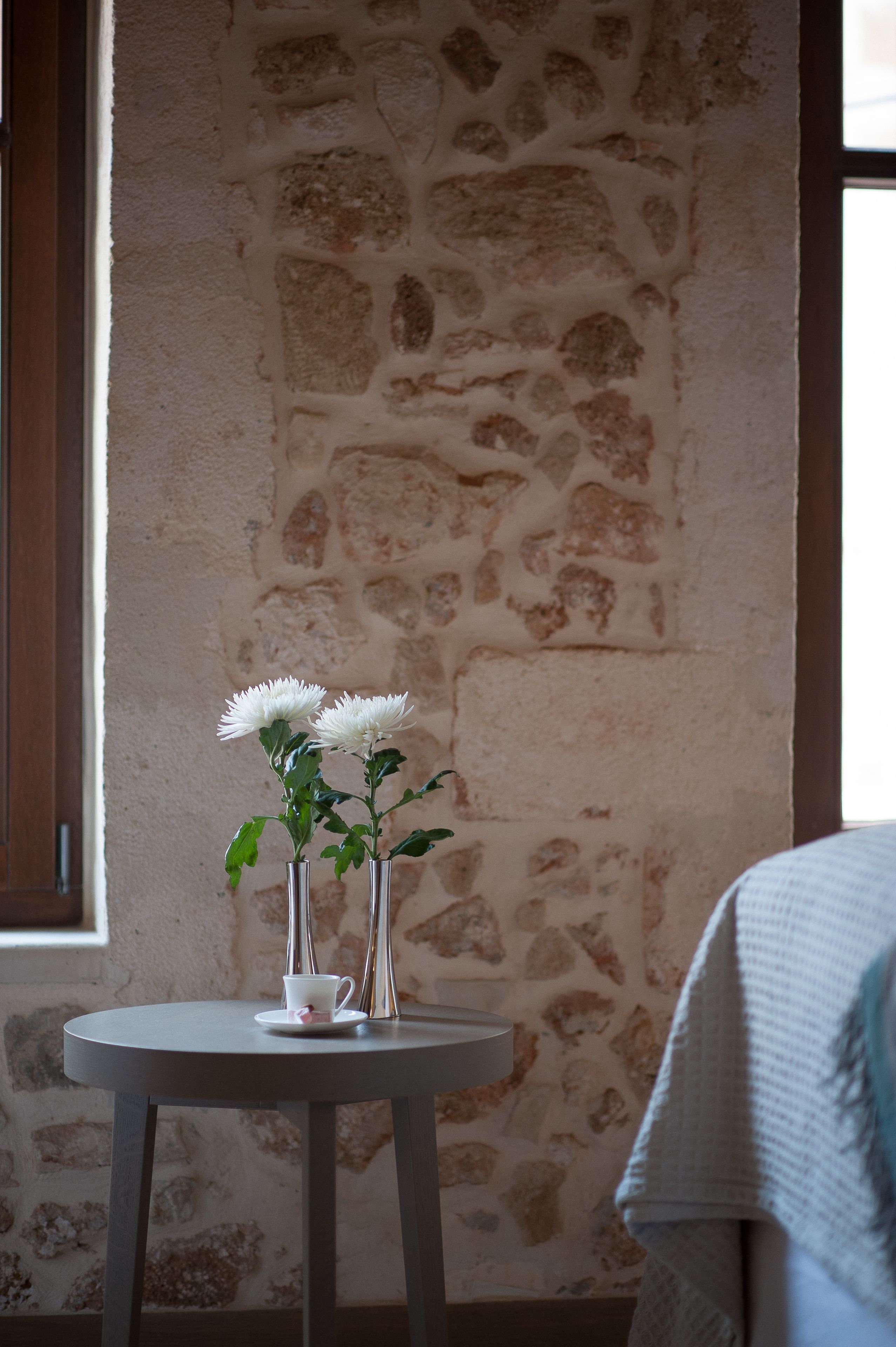 Un tavolo rotondo con due vasi bianchi colmi di fiori e una tazza bianca, posto di fronte a un muro in pietra con una finestra a sinistra.