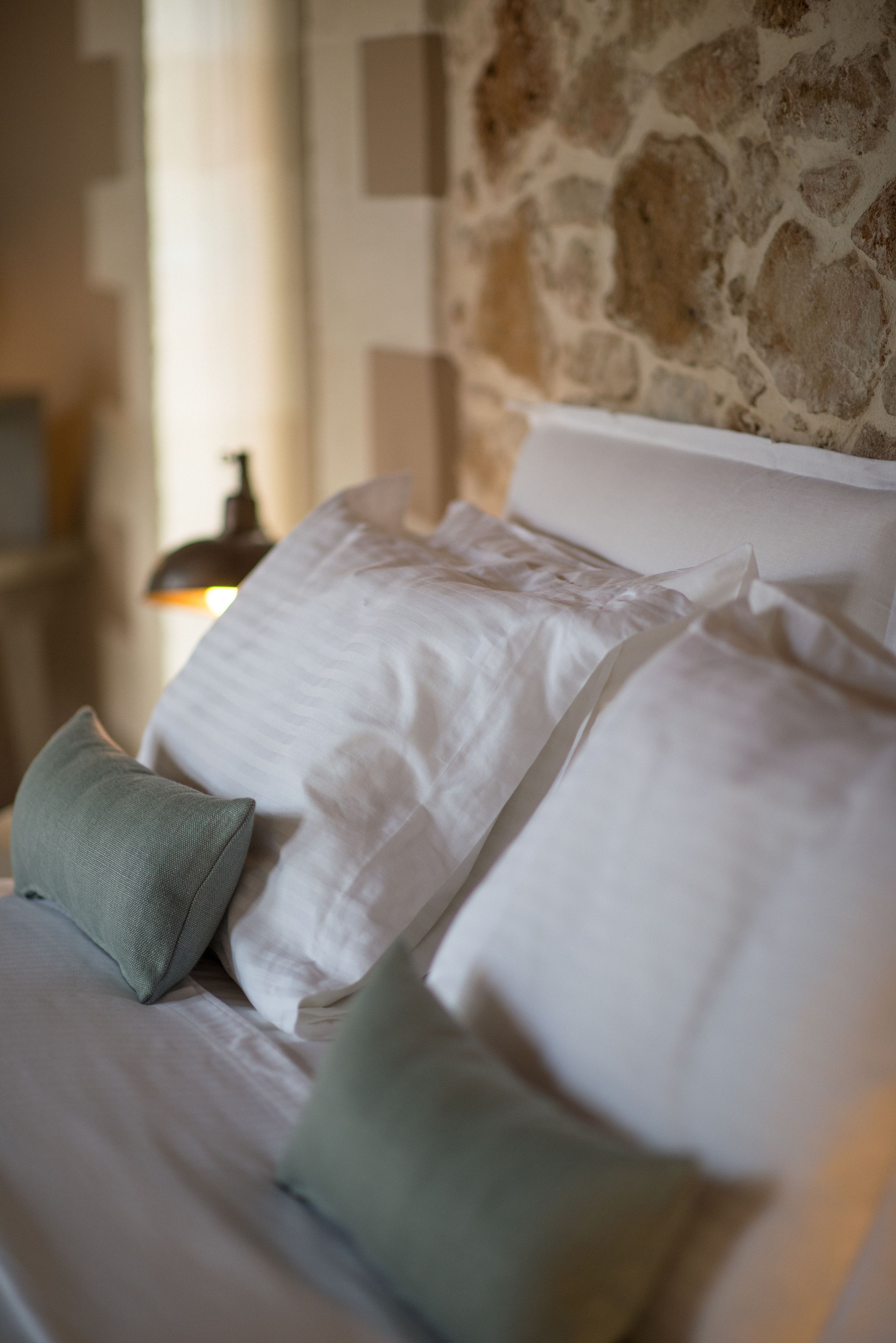 Un lit avec du linge de lit blanc et deux oreillers, un mur de pierre en arrière-plan et une lampe à l’arrière.
