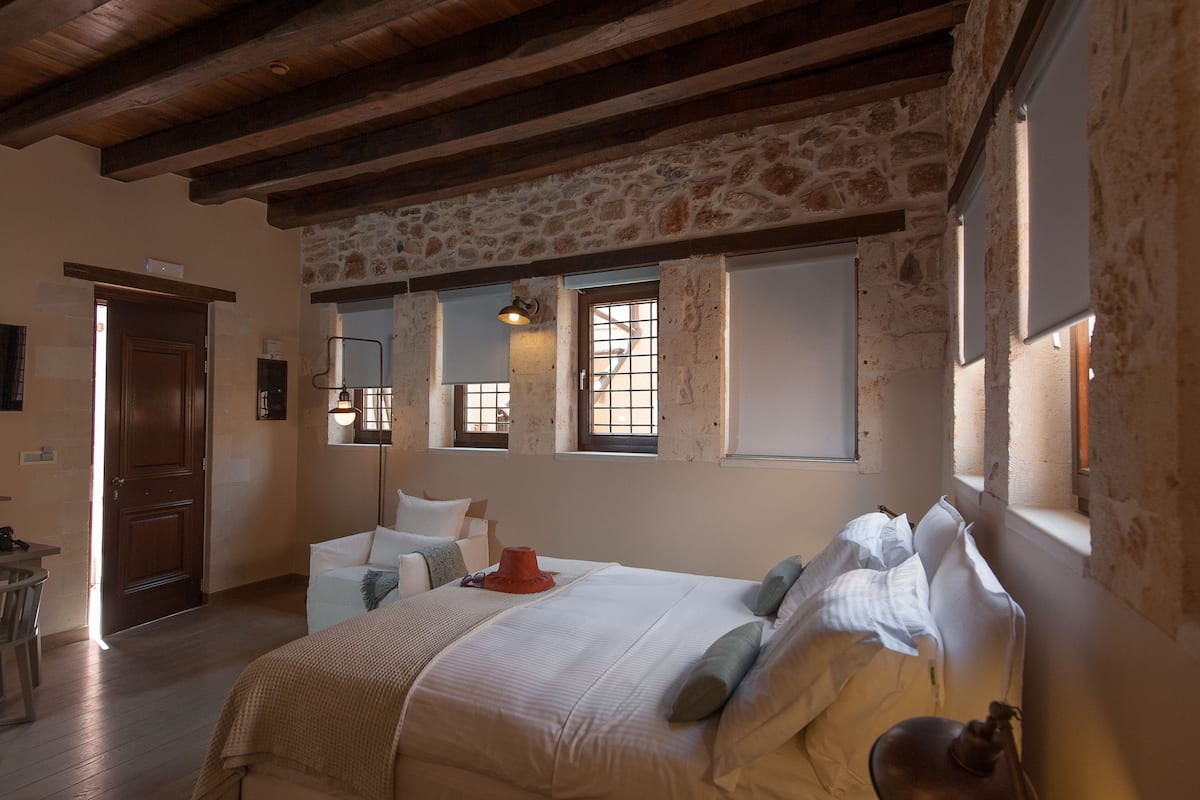 Une chambre avec un mur en pierre, des poutres en bois, un lit avec du linge de lit blanc, un chapeau rouge et un bureau avec une chaise.