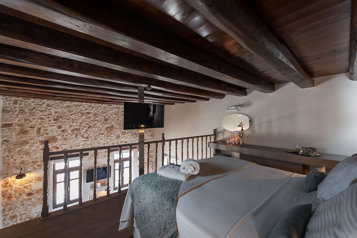 Une chambre avec un mur en pierre, des poutres en bois, une télévision, une table de chevet et une fenêtre.