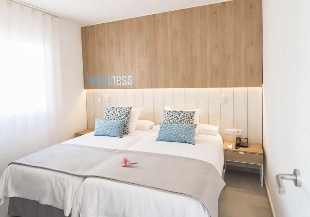 A hotel room with a wooden headboard, two beds with patterned pillows, a bedside table with a phone, and a wall-mounted light.