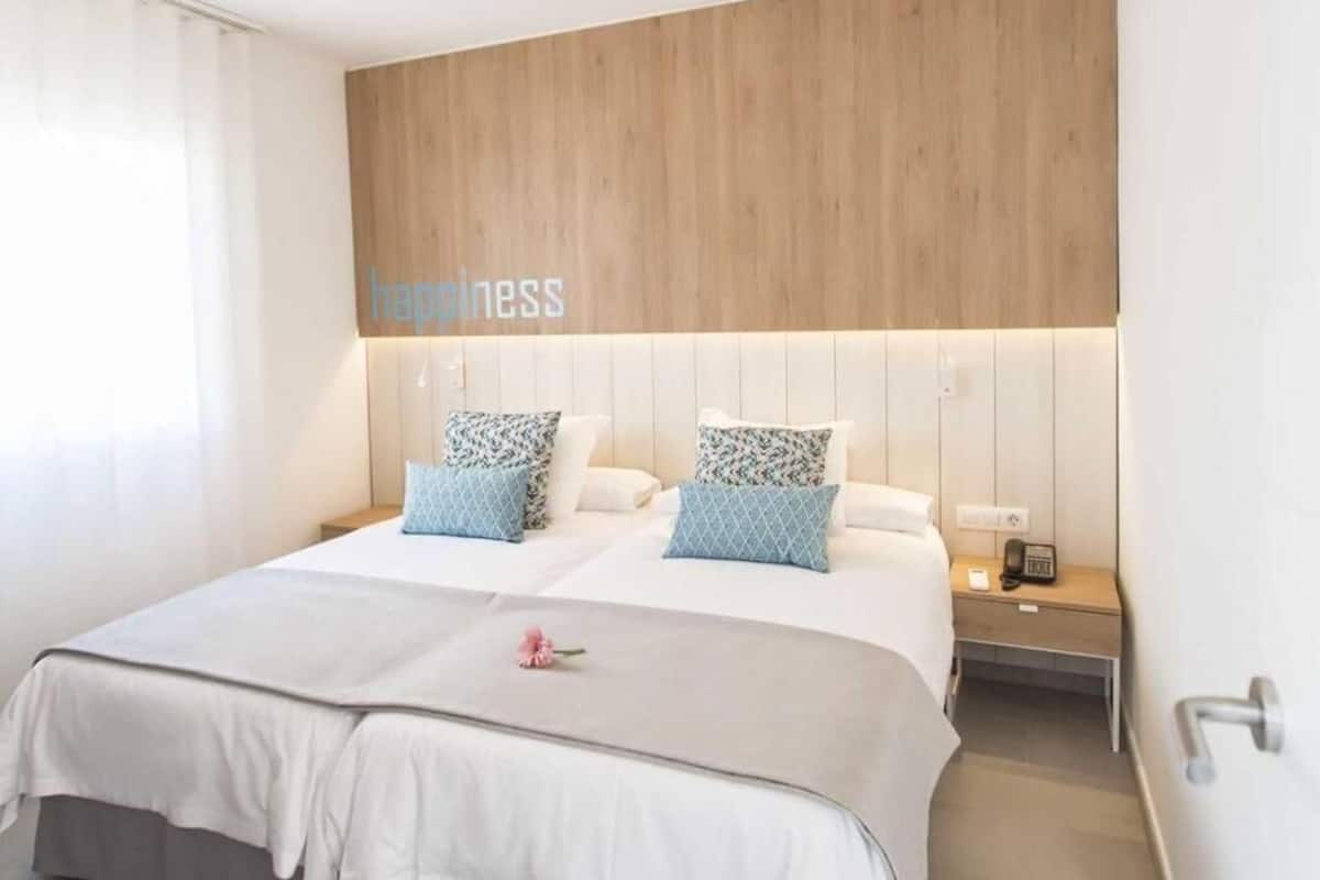 A hotel room with a wooden headboard, two beds with patterned pillows, a bedside table with a phone, and a wall-mounted light.