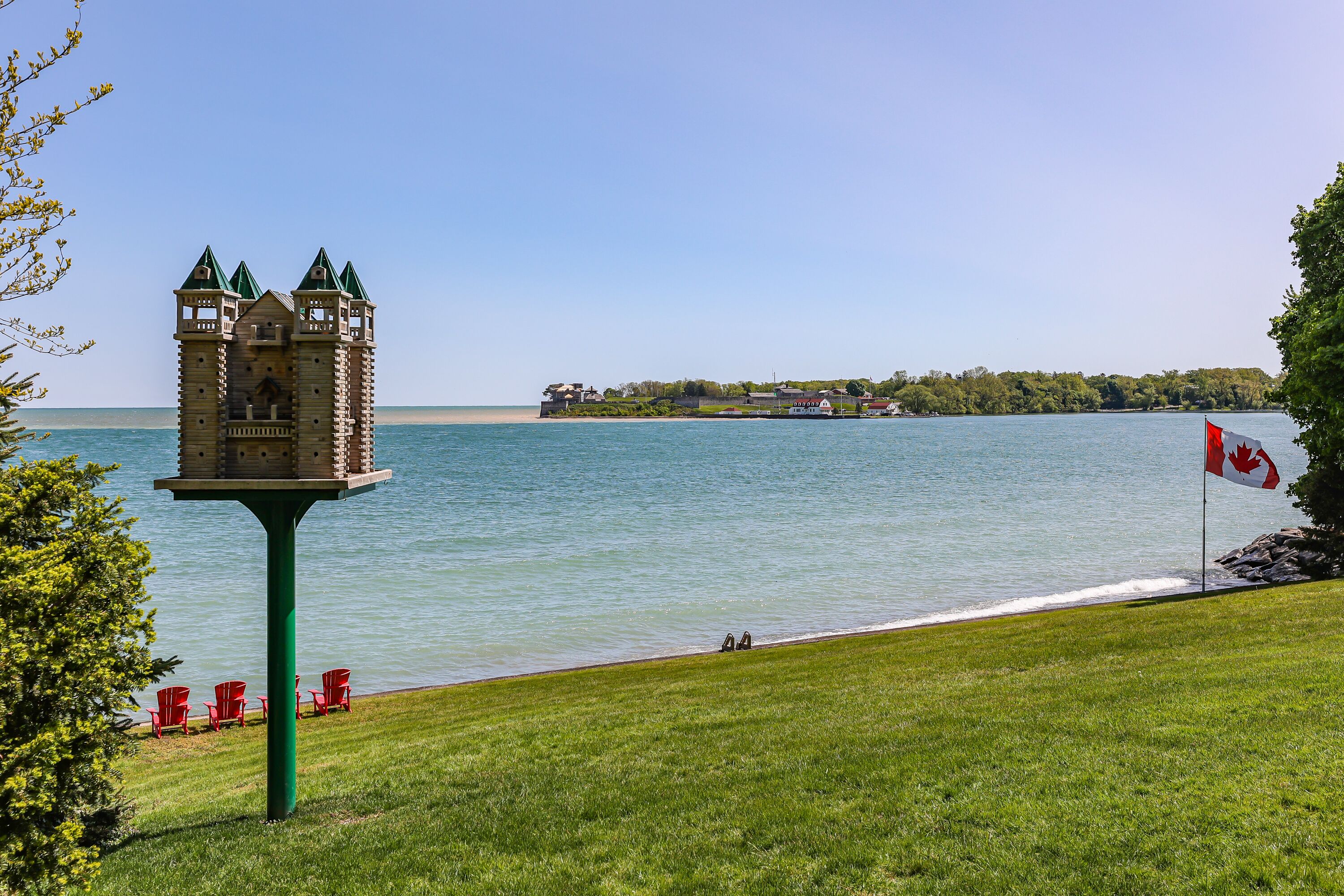 A lakeside view with a castle-like structure on a green pedestal, a Canadian flag, and red chairs along the shore.
