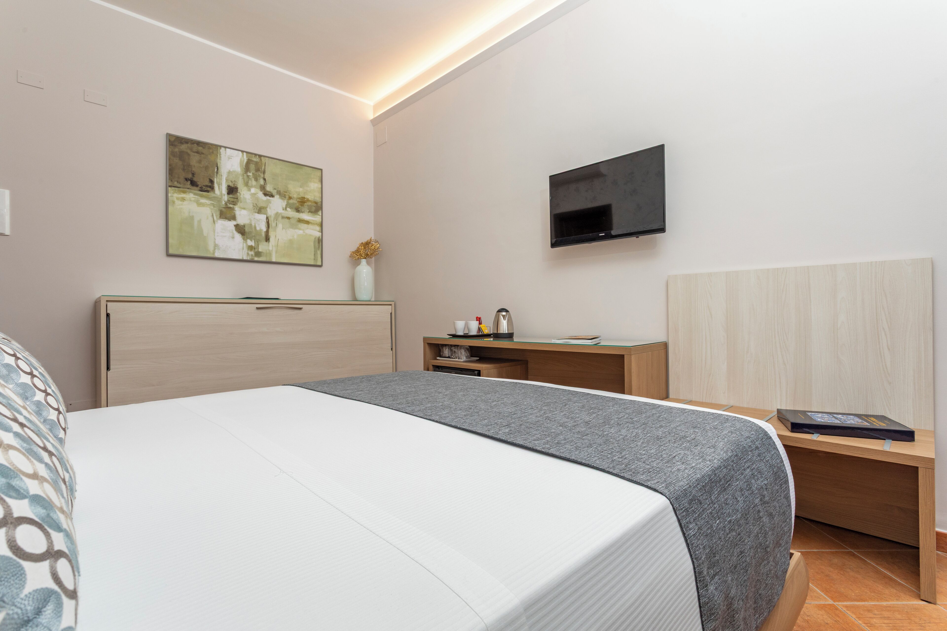 A modern hotel room with a bed, a desk, a TV, and a painting on the wall.