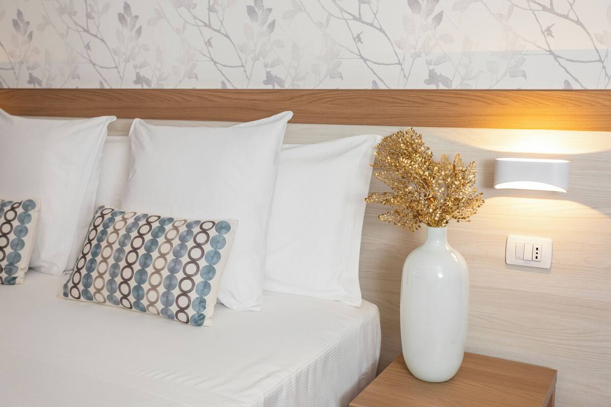 Ein ordentlich bezogenes Bett mit weißer Bettwäsche und gemusterten Kissen, eine weiße Vase mit getrockneten Blumen und eine wandmontierte Lampe.