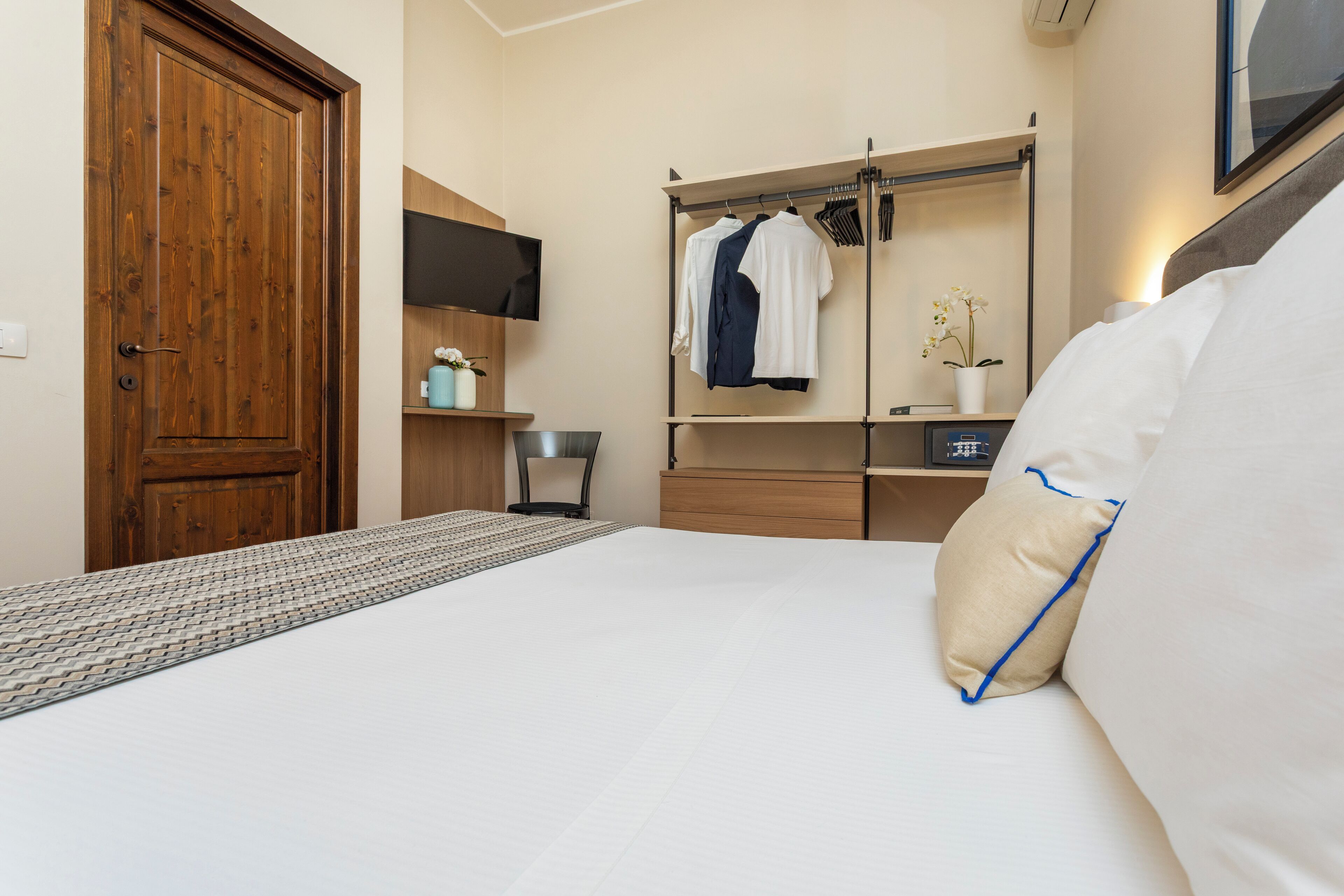 Een slaapkamer met een houten deur, een bed met wit beddengoed, een nachtkastje met een lamp, een spiegel en een kledingrek met hangende kleding.