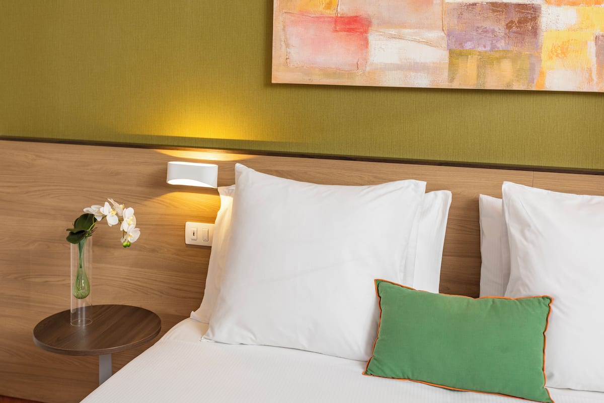 Ein Hotelzimmer mit Bett, weißen Kissen, einem grünen Kissen, einer Vase mit Blumen, einem Nachttisch und einer Wandleuchte.