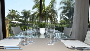 Une table à manger est dressée pour six personnes, avec de la vaisselle transparente, des serviettes blanches et des couverts en métal. La table est placée sur un balcon d’où l’on peut admirer des palmiers et un ciel dégagé.