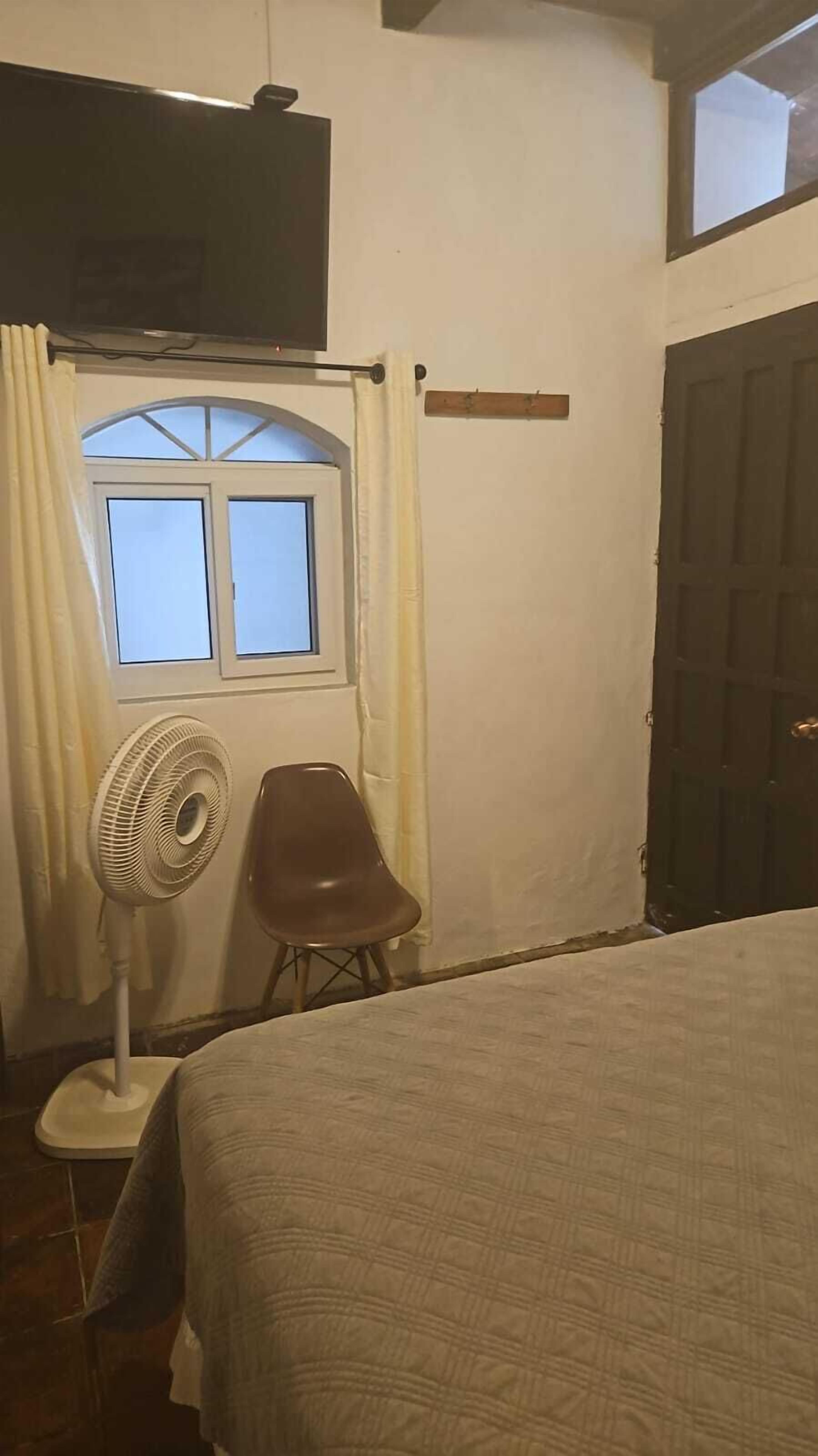 Una habitación con cama, una silla, un ventilador de pie, una ventana con cortinas y una puerta.