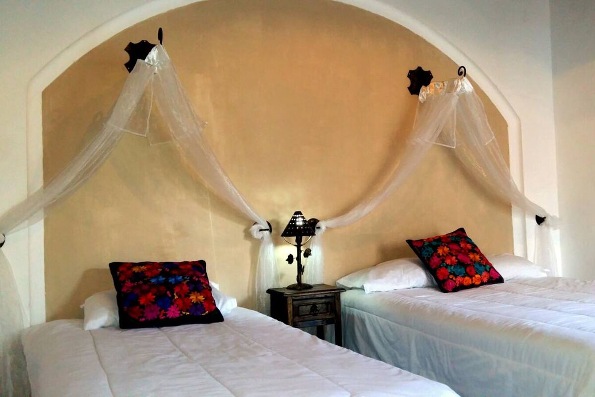 A room with two beds, each with a white bedspread and a colorful floral pillow. There is a nightstand with a lamp between the beds.