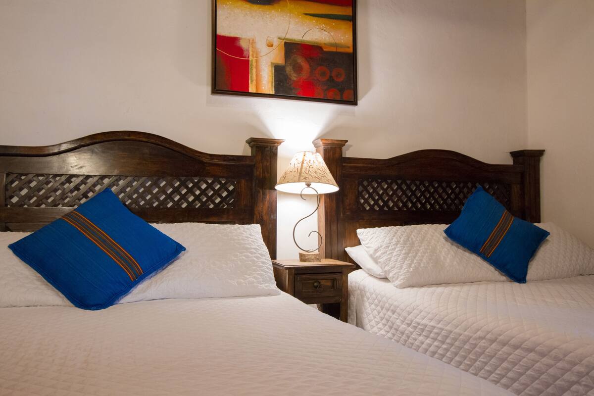 A hotel room with two beds, each with white bedding and blue pillows. There is a wooden headboard with a lattice design, and a lamp on a bedside table. A colorful abstract painting hangs on the wall above the beds.