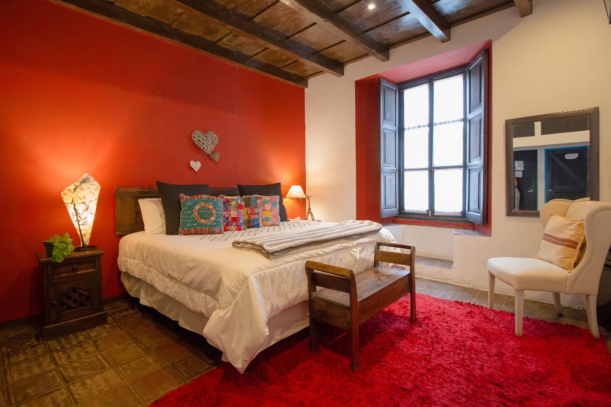 A bedroom with a red carpet, a wooden bench, a white bed with patterned pillows, a nightstand with a lamp, and a window with a view.