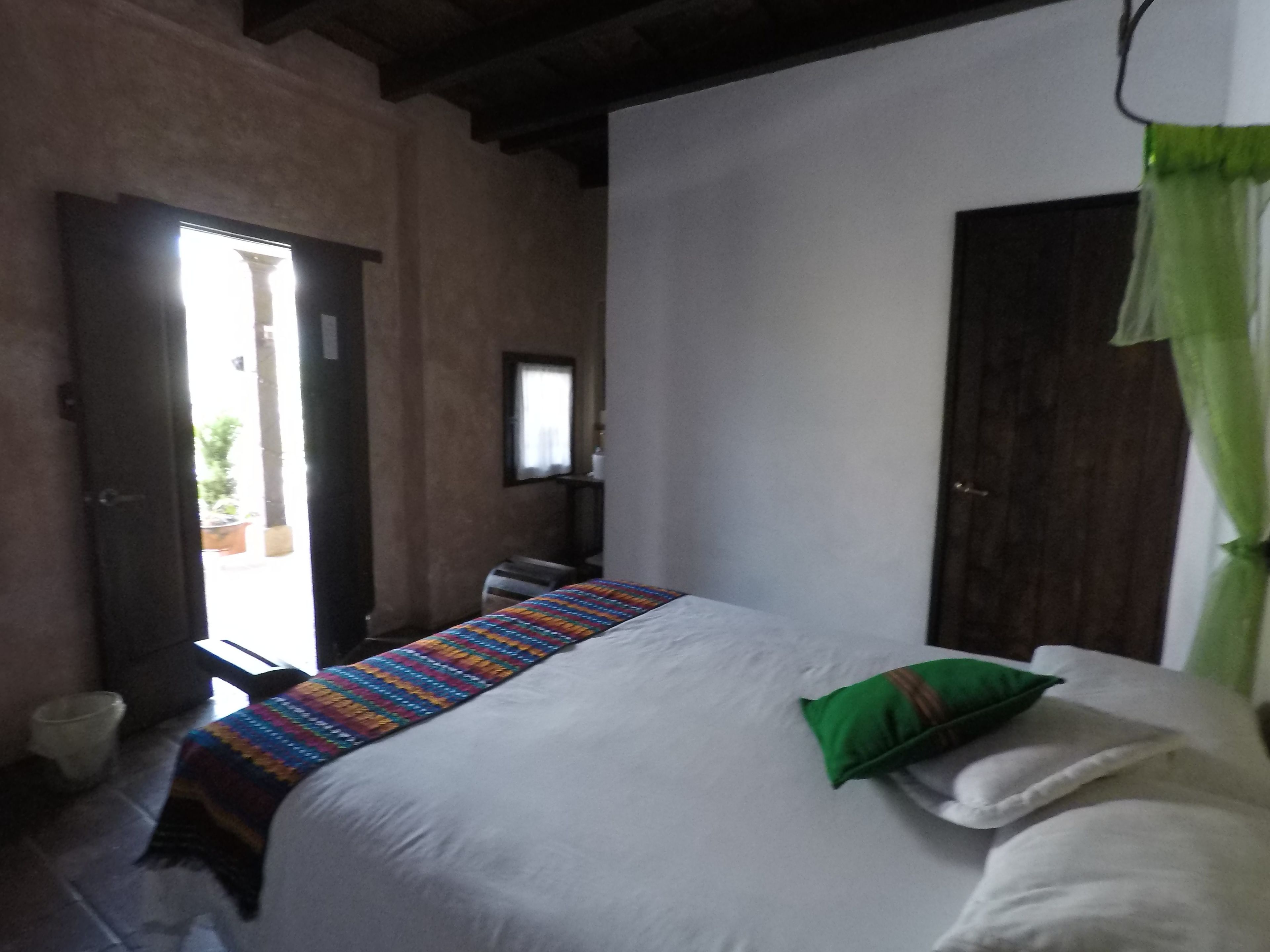 Un dormitorio con una cama, un almohadón verde, una colcha colorida, una puerta de madera y una ventana con cortinas.
