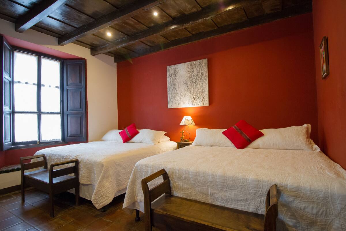 Two single beds with white linens and red pillows, a wooden chair, a window with a view, and a wall-mounted lamp.