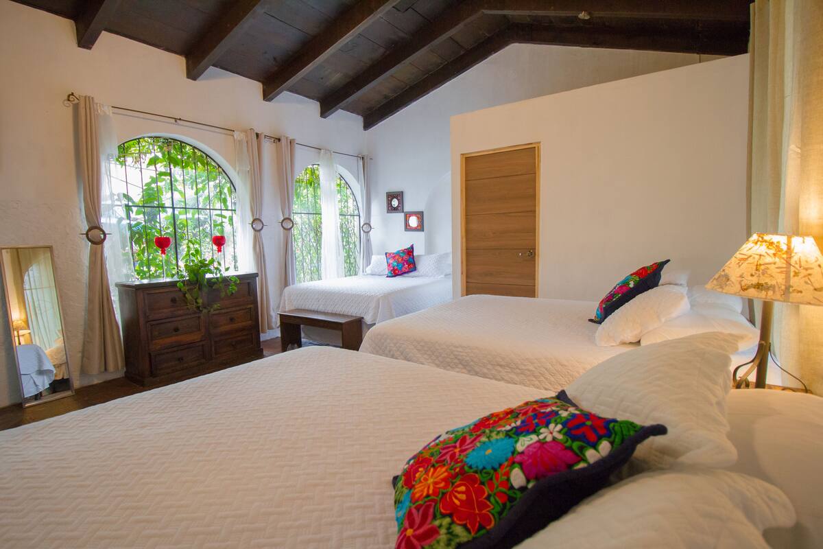 A bedroom with two beds, a wooden wardrobe, a dresser, a window with curtains, and a lamp.