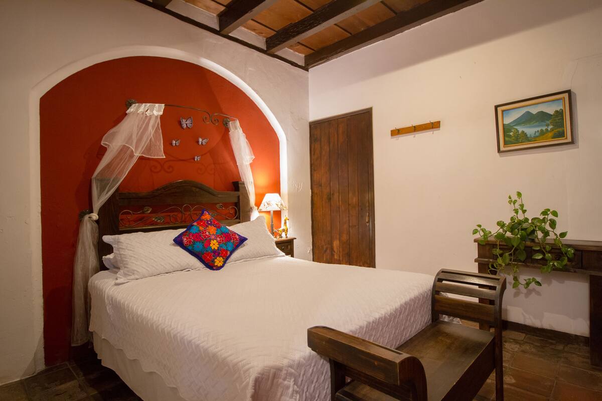 A bed with a canopy, a wooden bench, a nightstand with a lamp, and a painting on the wall.