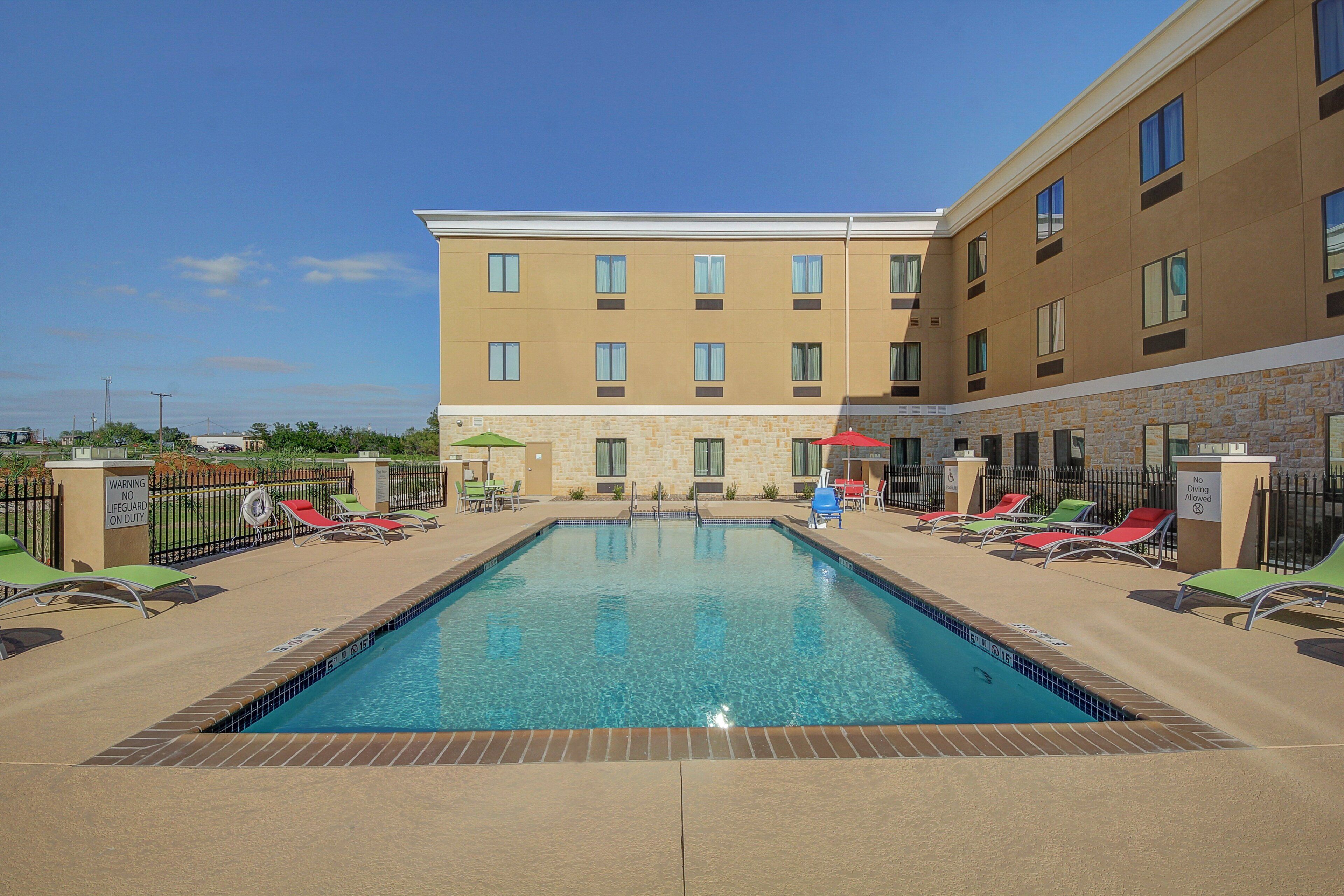A hotel pool area with a clear blue pool, sun loungers, and a building with multiple windows.