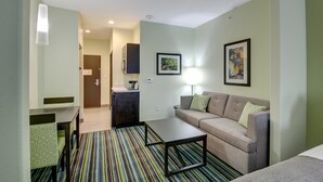 A compact living space with a sofa, a coffee table, a striped rug, a dining table with chairs, a kitchen area, and artwork on the walls.