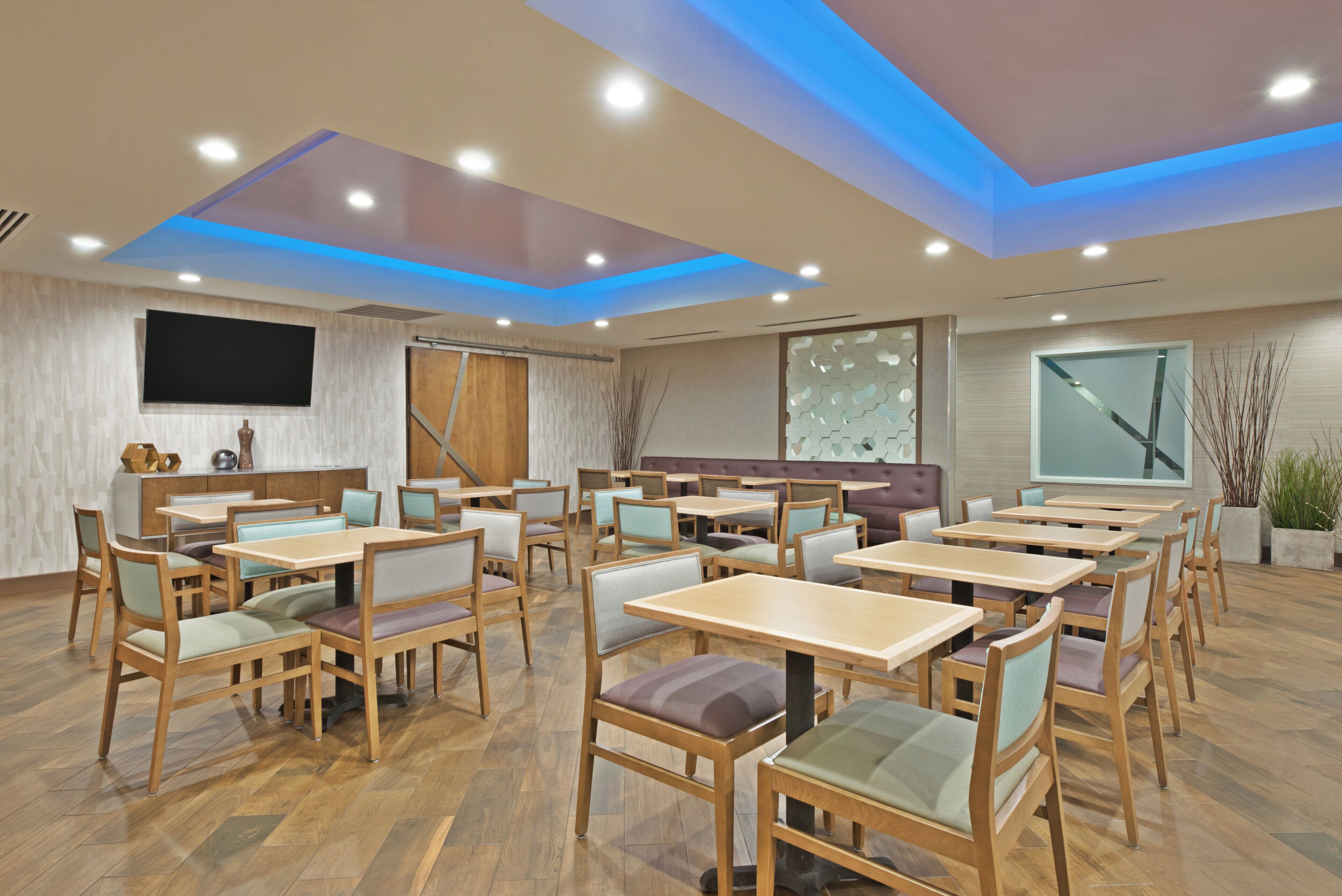 A modern dining area with wooden tables and chairs, recessed ceiling lighting, and a large flat-screen TV mounted on the wall.