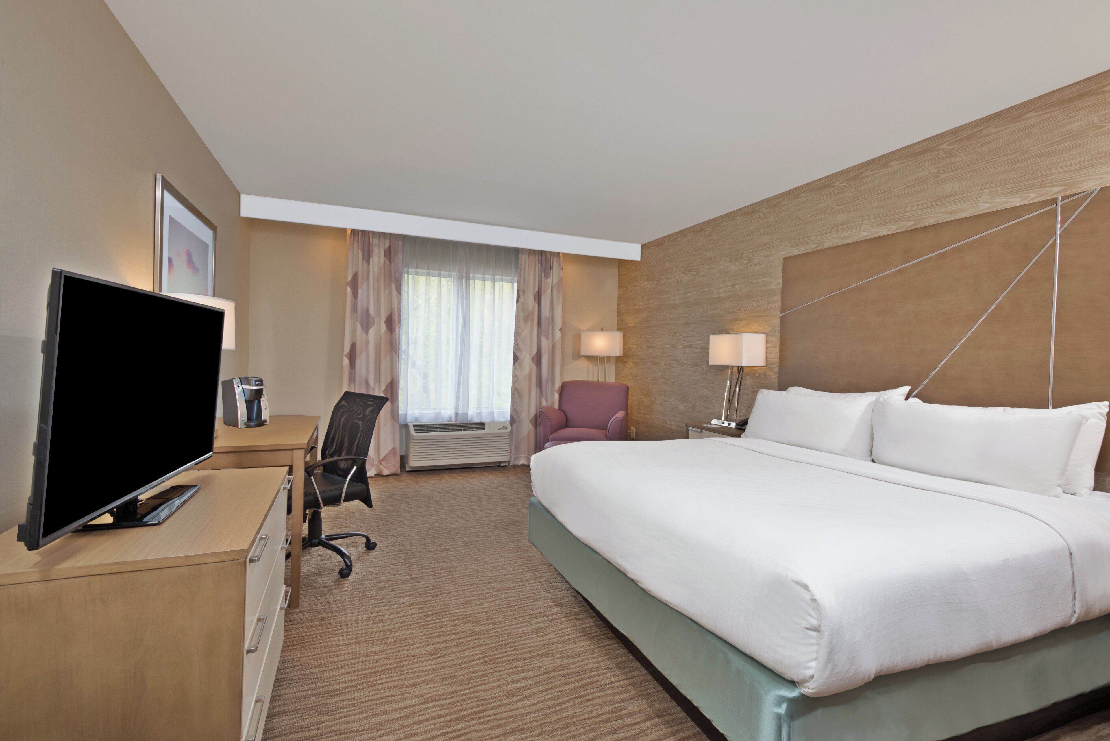 A hotel room with a bed, a desk with a television, a chair, a desk lamp, and a window with curtains.