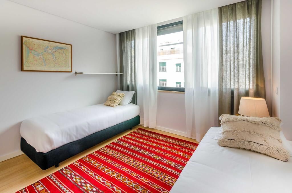 A hotel room with a bed, a red patterned rug, a map on the wall, and a window with curtains.
