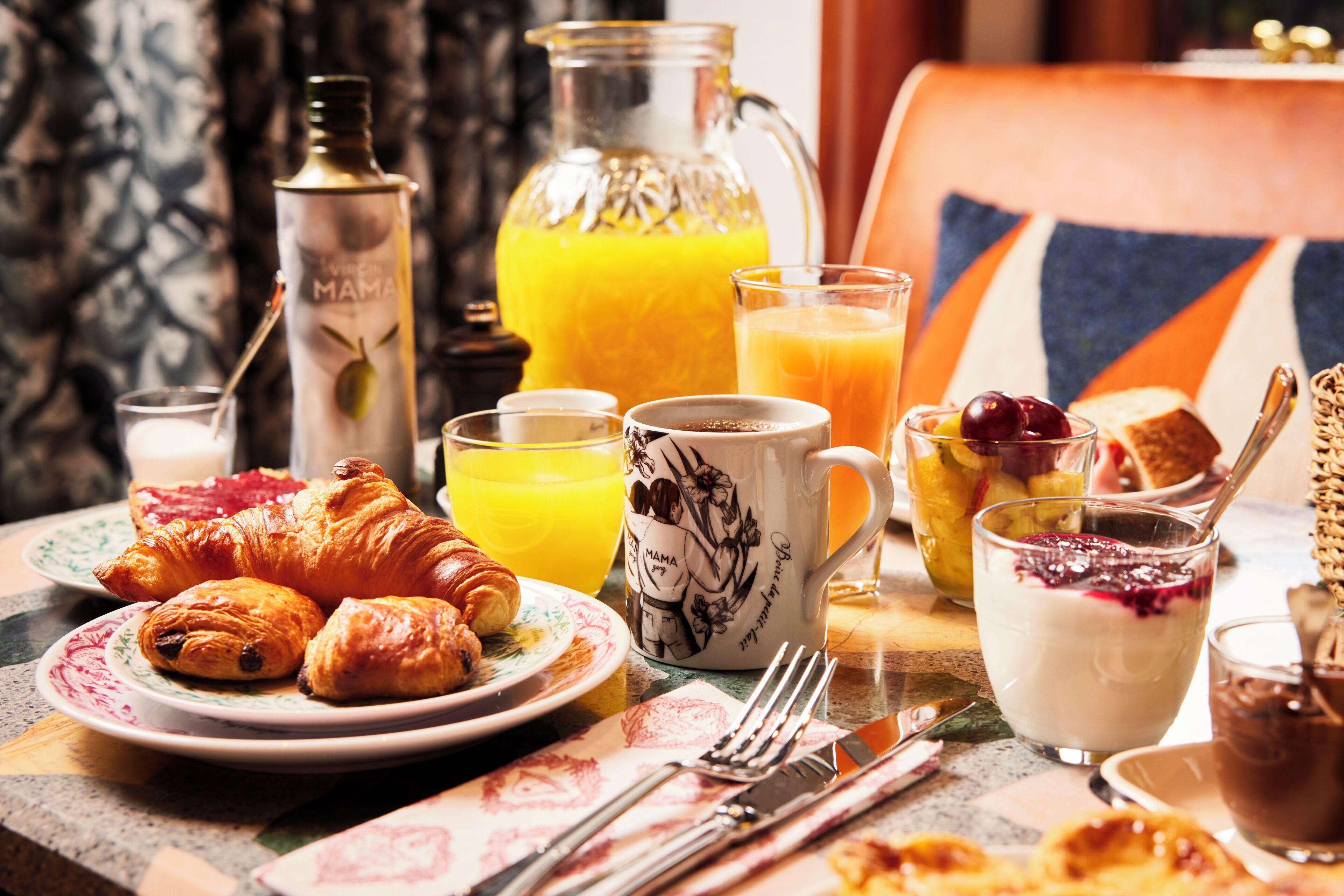 Ett frukostbord med croissanter, apelsinjuice, en kanna apelsinjuice, en mugg, en tallrik med rostat bröd och sylt samt ett glas yoghurt med bär.