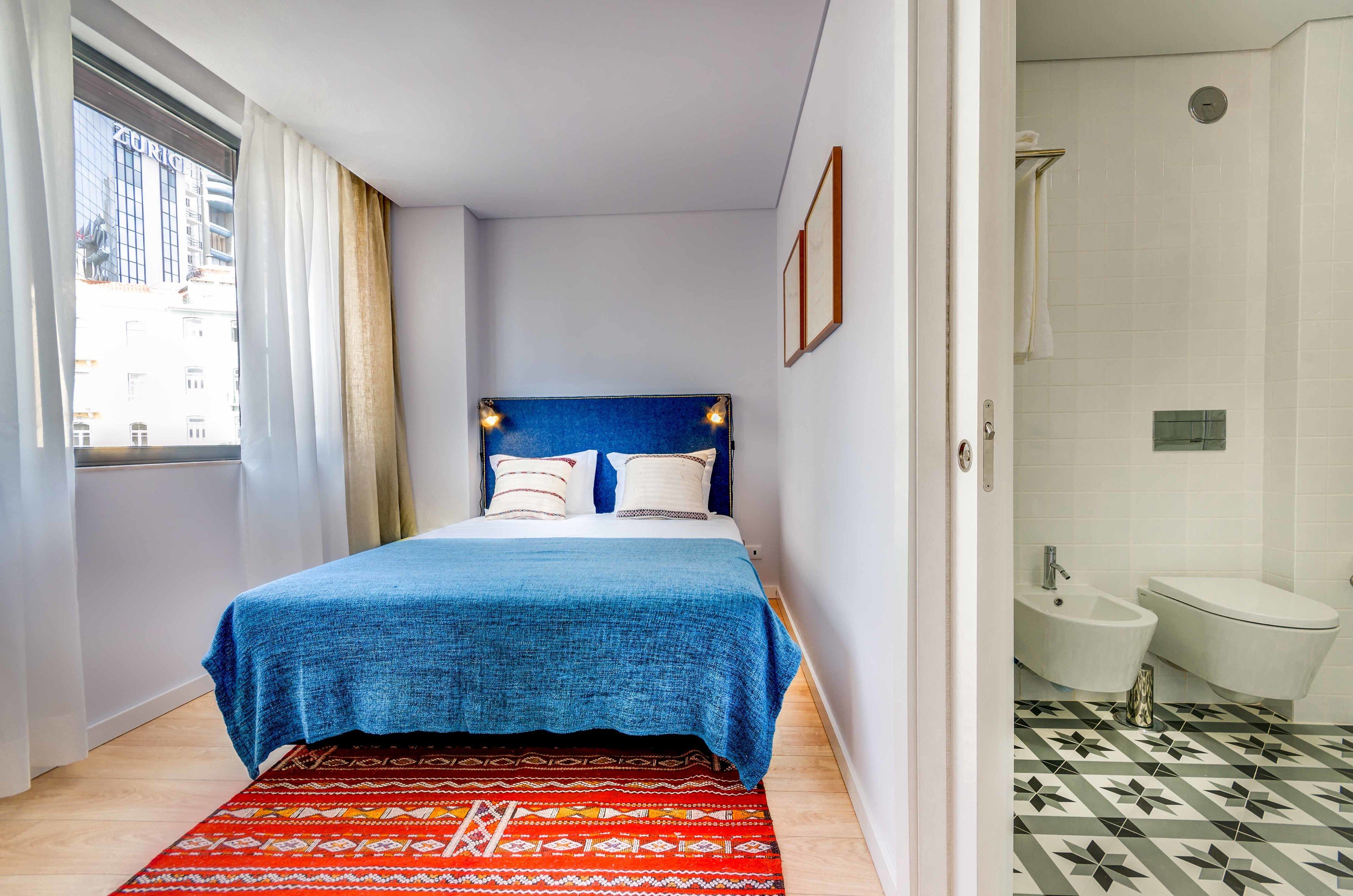 A bedroom with a bed, a window with curtains, a bathroom with a sink and toilet, and patterned flooring.