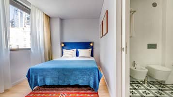 A bedroom with a bed, a window with curtains, a bathroom with a sink and toilet, and patterned flooring.
