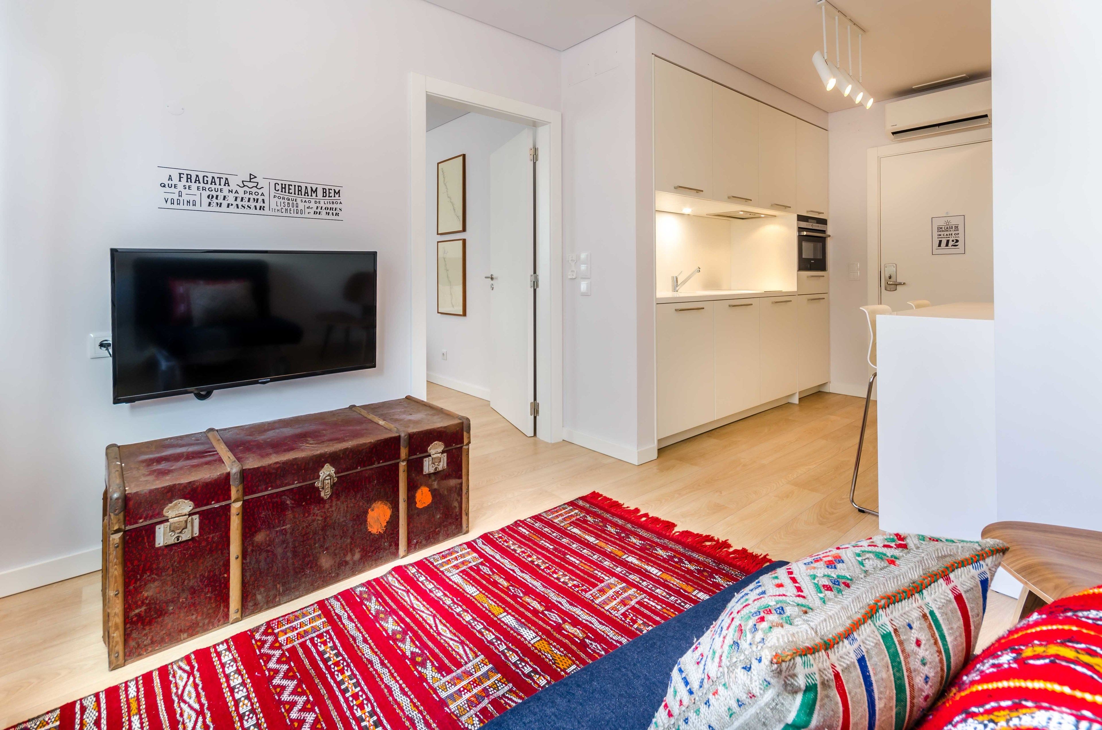 A modern living room with a flat-screen TV, a vintage suitcase, a red patterned rug, and a sofa with colorful cushions.