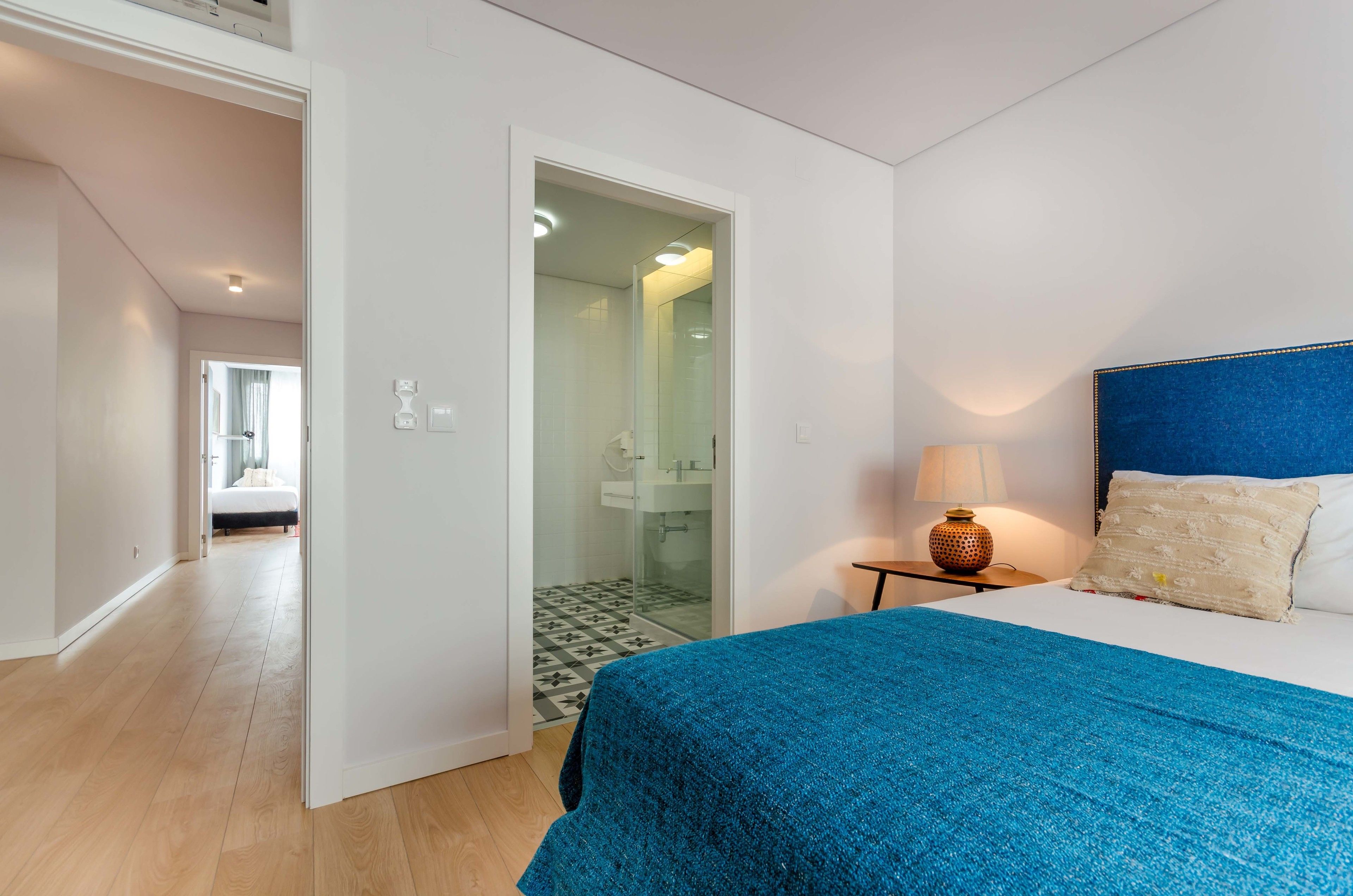 A bedroom with a bed, a nightstand with a lamp, and a bathroom visible through an open door.