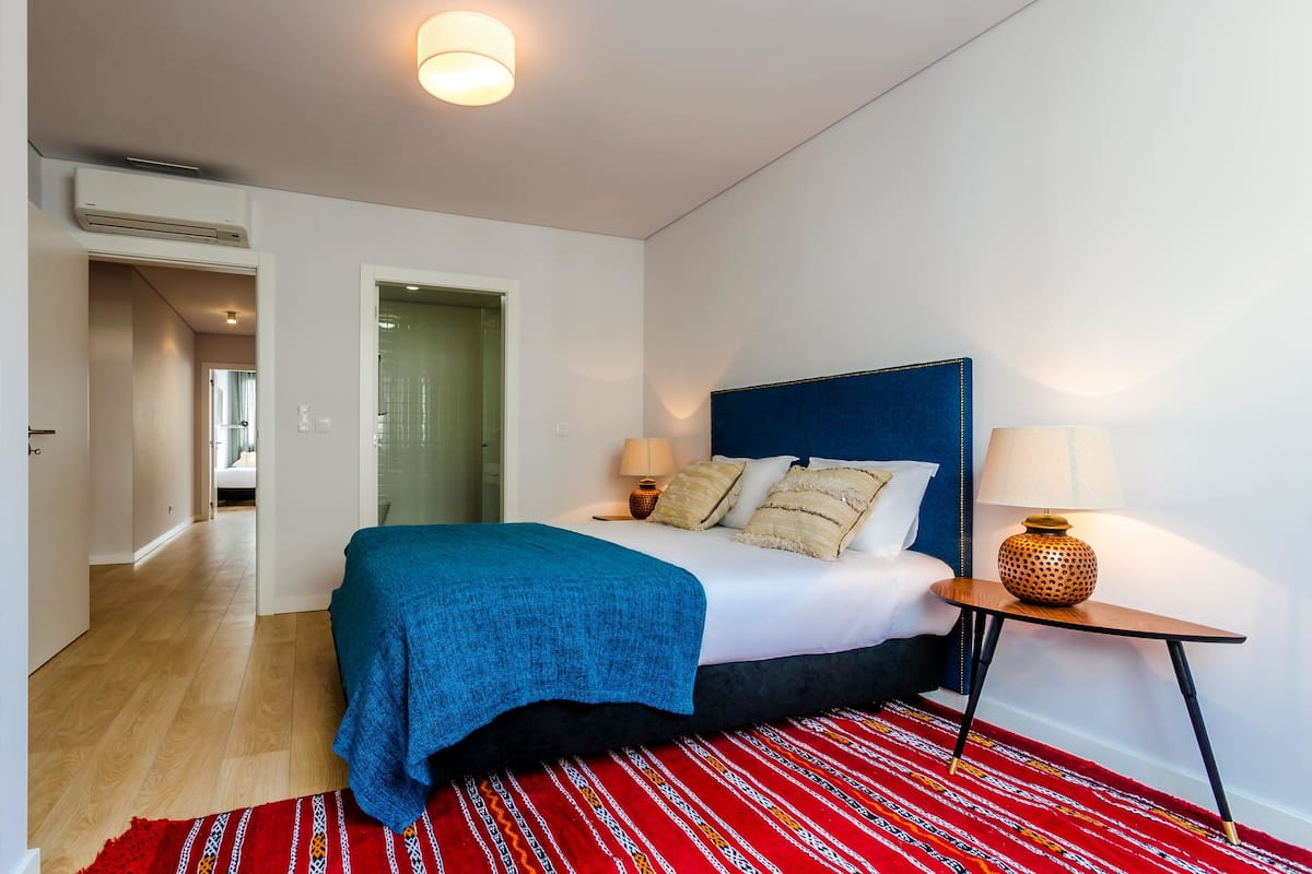 Een slaapkamer met een bed, nachtlampjes, een rood geweven vloerkleed en een blauw hoofdeinde.
