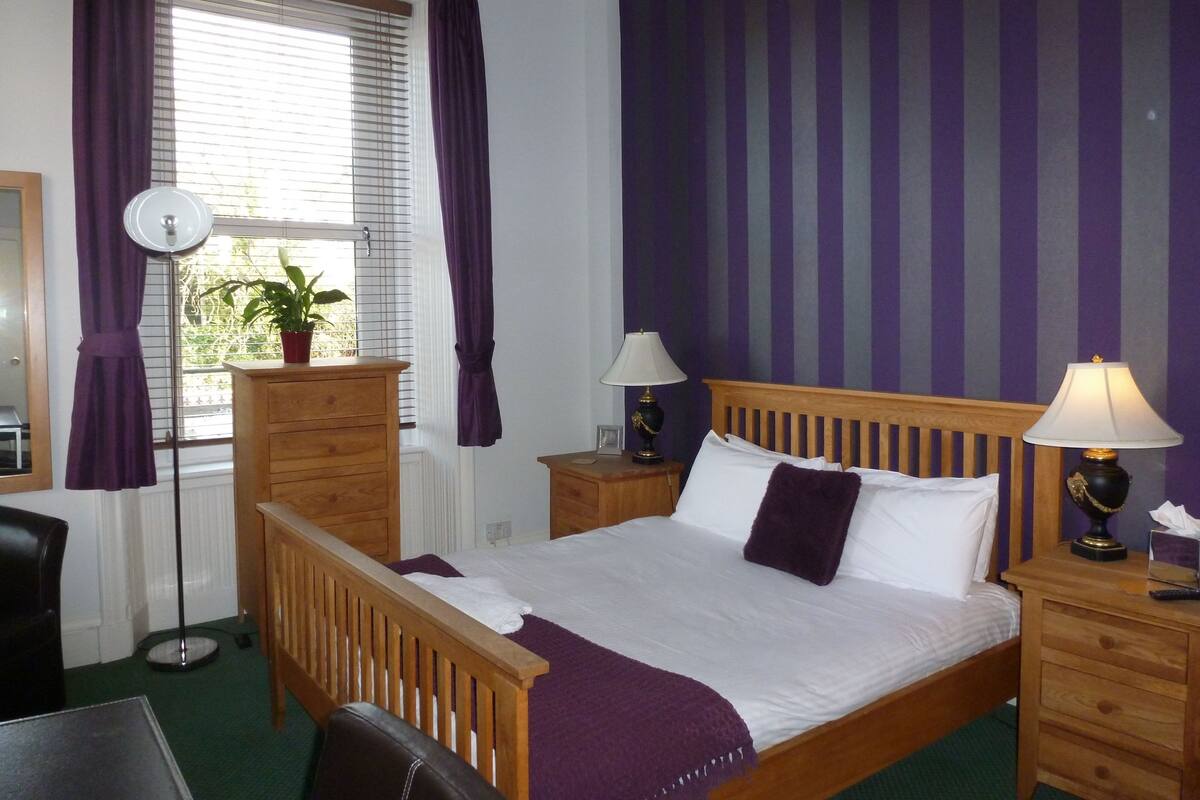 A bedroom with a wooden bed, two bedside tables with lamps, a dresser, a mirror, and a window with curtains.