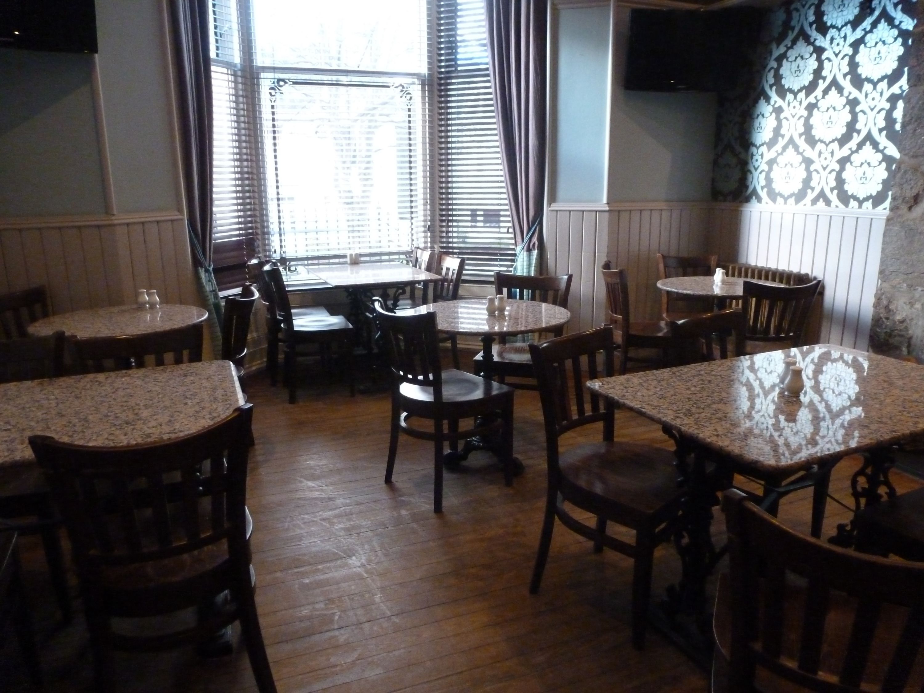 A restaurant interior with wooden tables and chairs, a large window with blinds, and a patterned wall.