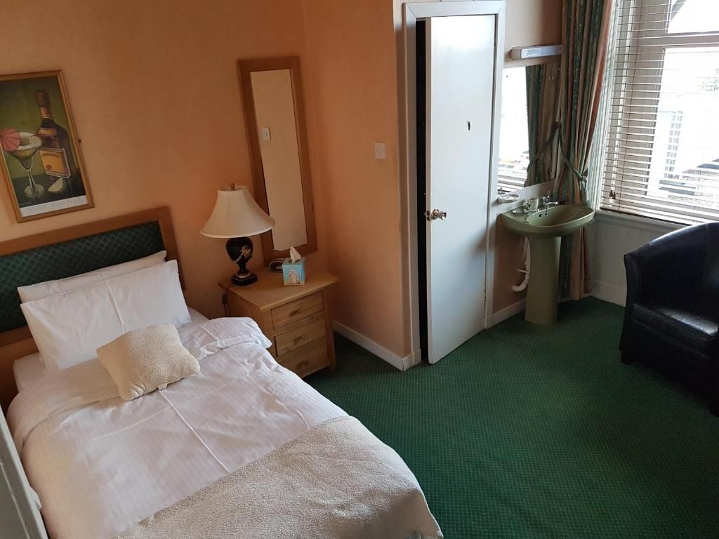 A hotel room with a bed, a nightstand, a lamp, a mirror, a bathroom area, and a window with blinds.