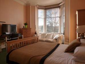 A hotel room with a bed, a television, a desk with a computer, a chair, and a window with blinds.