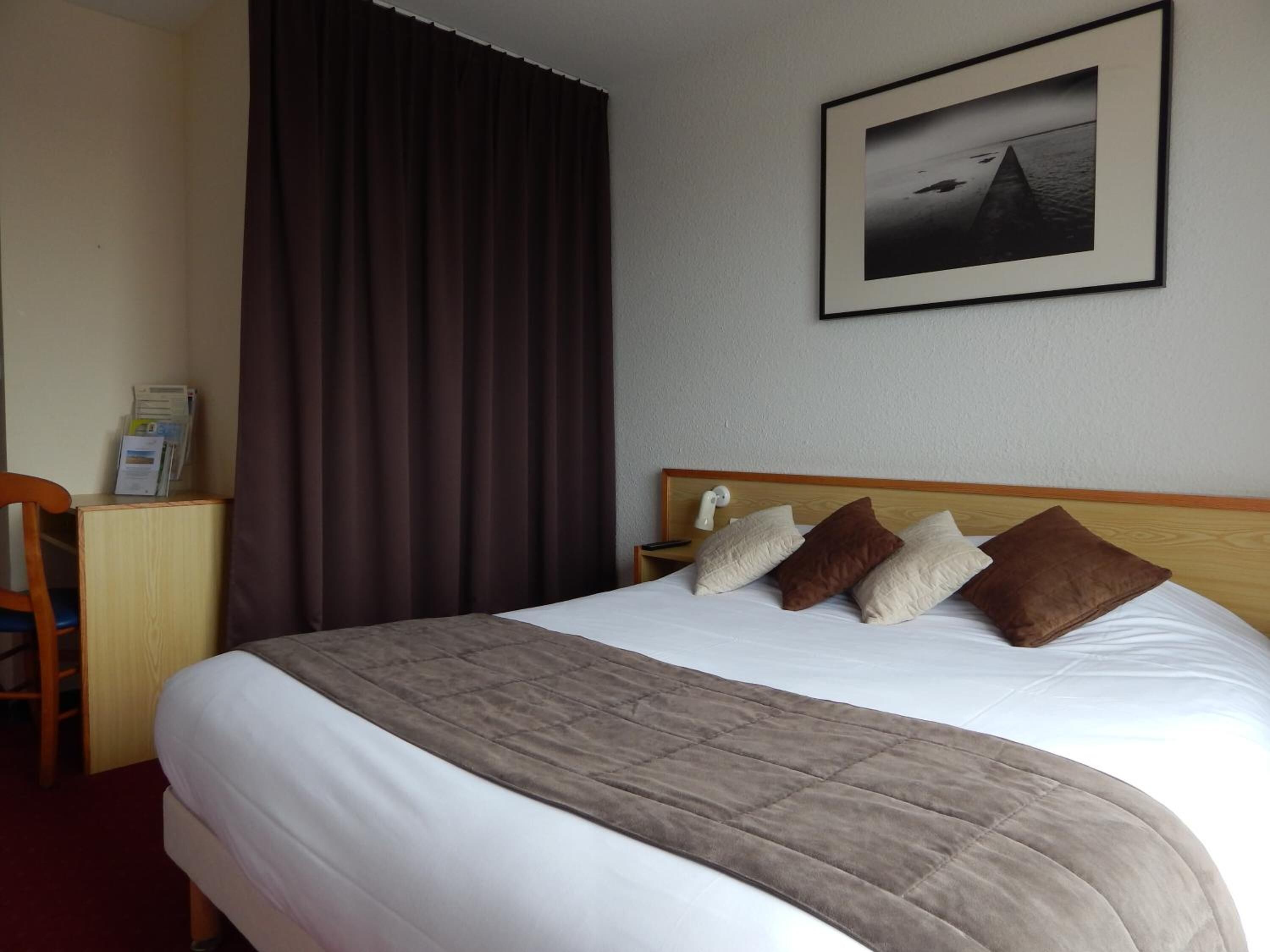 Hotellihuone, jossa on suuri sänky, puinen pääty, ruskea peitto ja seinällä oleva kehystetty kuva.