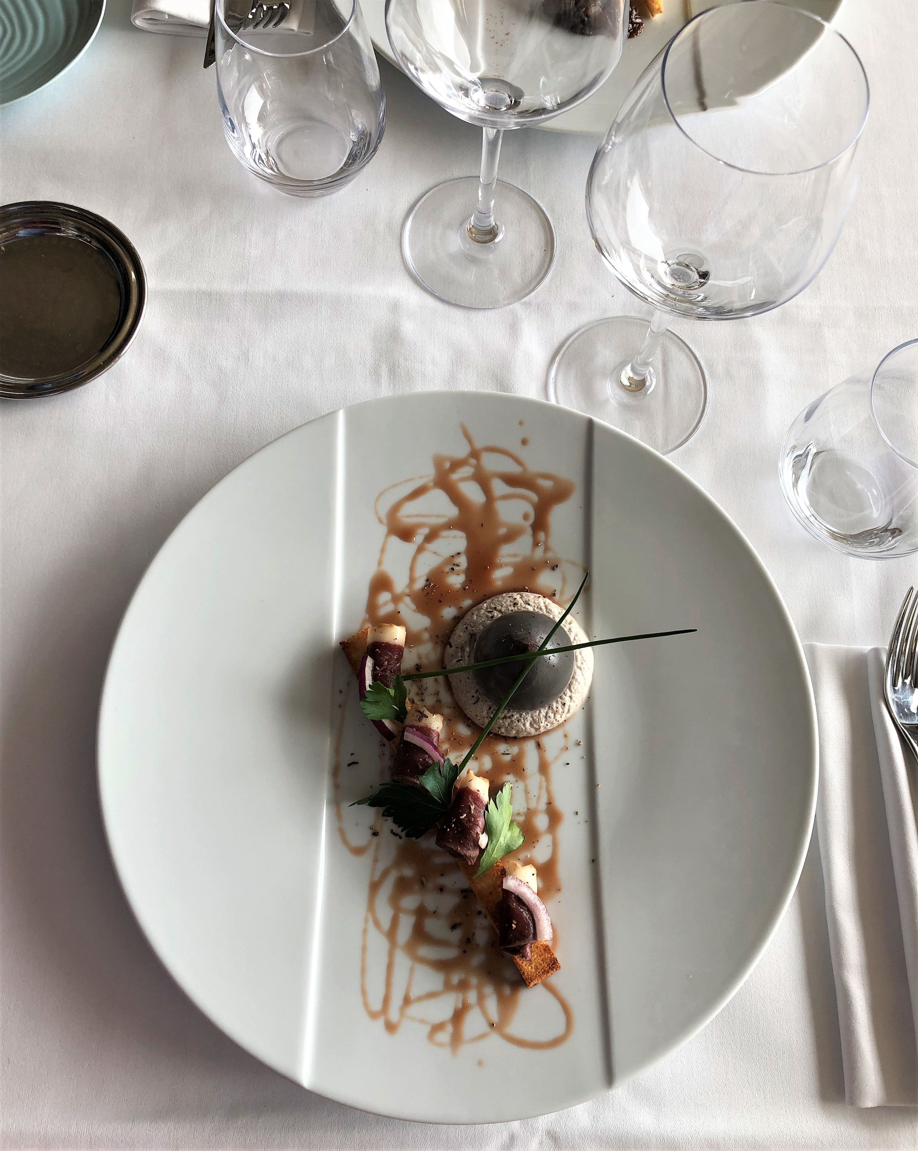 A white plate with a dessert, surrounded by empty wine glasses and a fork on a tablecloth.