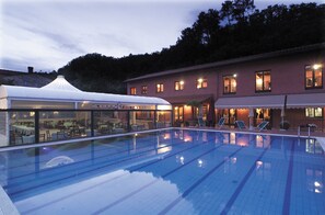 En swimmingpool med liggestole, et overdækket spiseområde og en bygning med flere vinduer og lys.