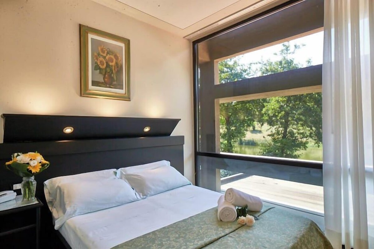 Quarto de hotel com cama, mesa de cabeceira, um vaso de flores, um quadro e uma janela ampla com vista para o verde.