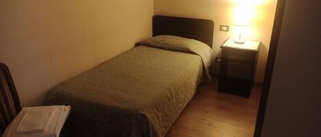 A small, single-bed room with a wooden floor, a bedside table with a lamp, and a chair with a folded towel.