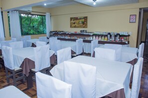 A dining area with tables covered in white tablecloths, chairs with brown seat covers, a buffet setup with various food items, and a painting on the wall.