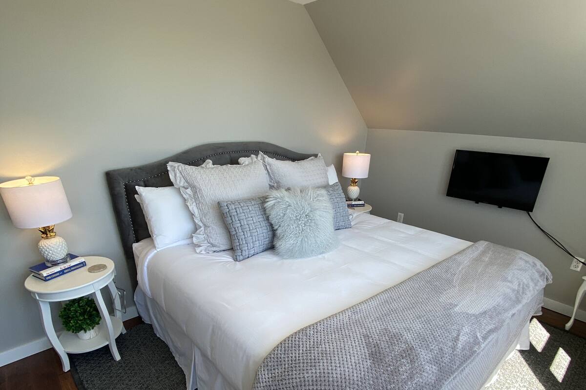 A neatly arranged bedroom with a large bed, two bedside tables with lamps, a flat-screen TV mounted on the wall, and a grey rug on the wooden floor.