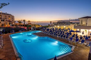 Une piscine entourée de bâtiments, dotée de chaises longues et de parasols, et offrant une vue sur la mer.