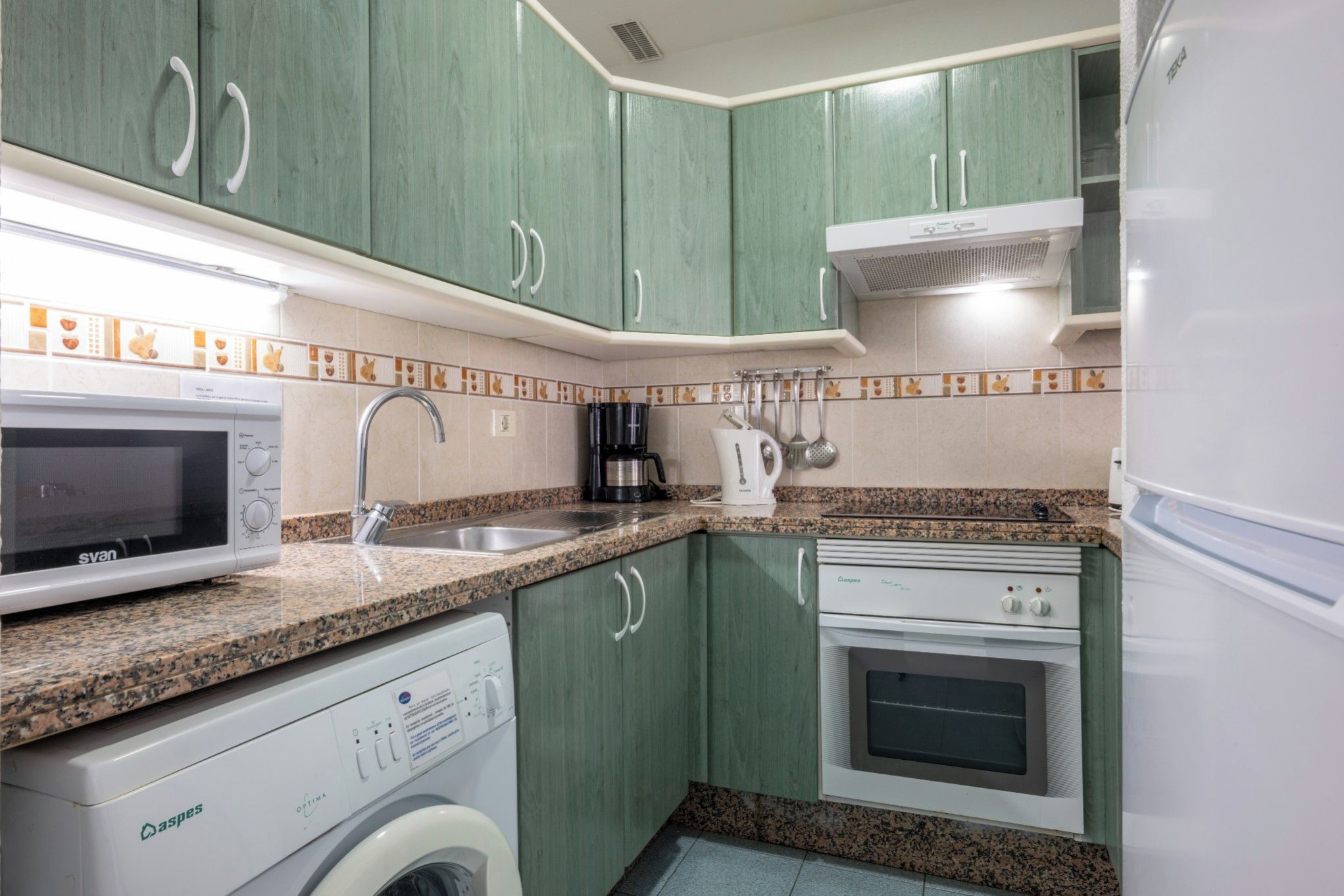 A compact kitchen with green cabinets, a microwave, a coffee maker, a washing machine, and a built-in oven.