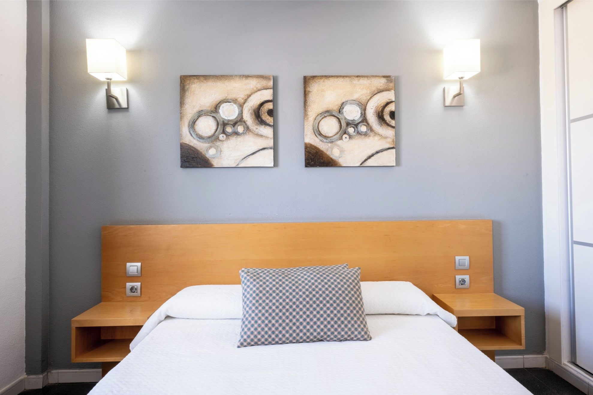 Een hotelkamer met een bed, twee wandlampen en twee abstracte schilderijen.