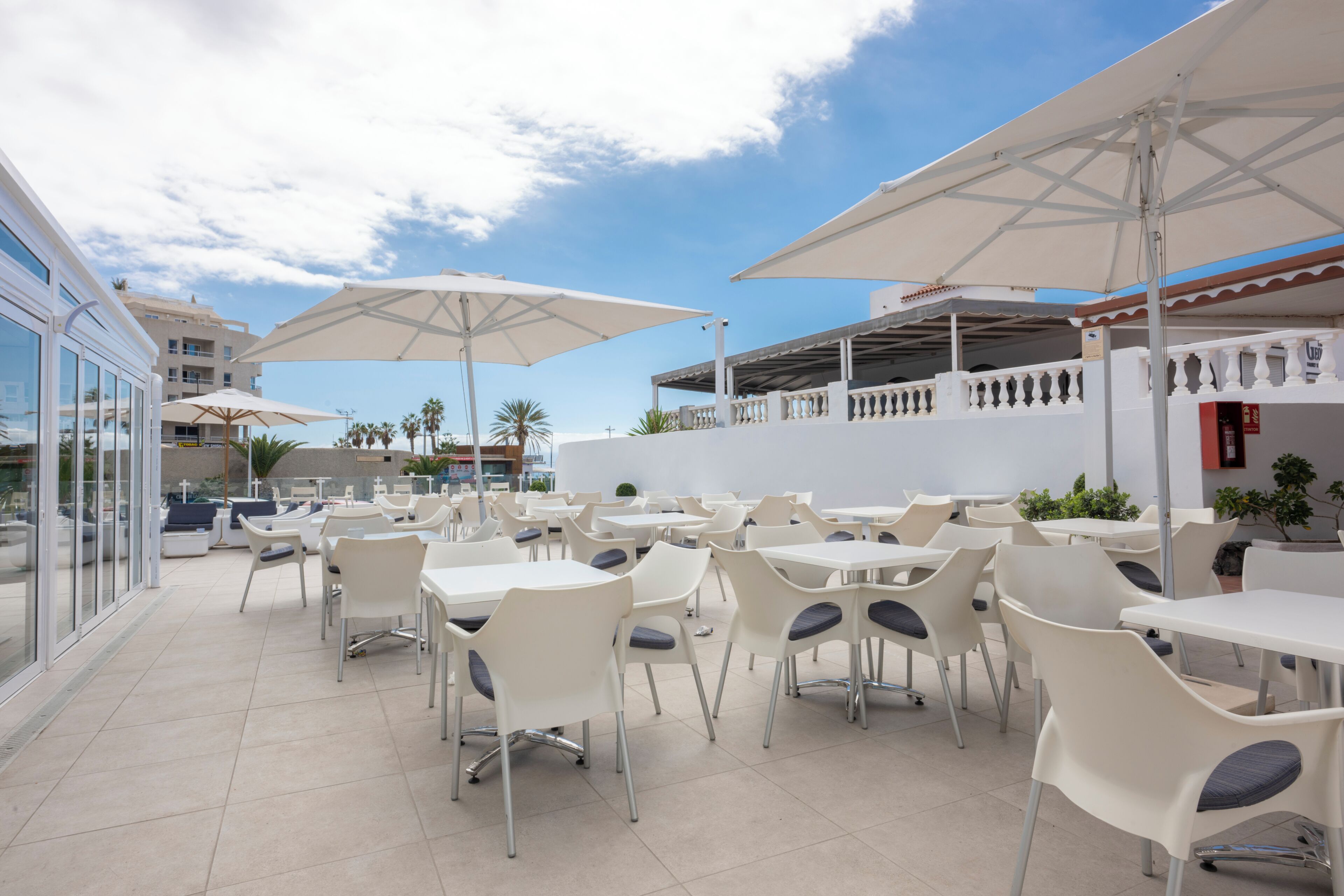 An outdoor dining area with white tables and chairs, umbrellas, and a view of the sea and buildings.